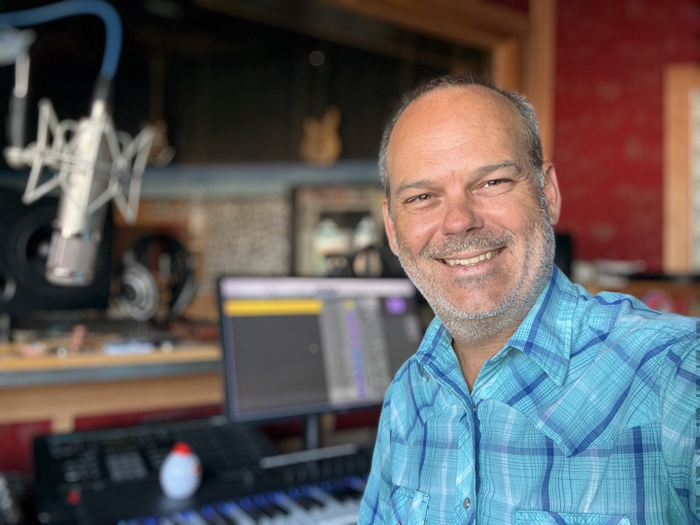 Portrait of Jamey Warren in a blue plaid shirt, smiling, with a studio mic and keyboard behind him.